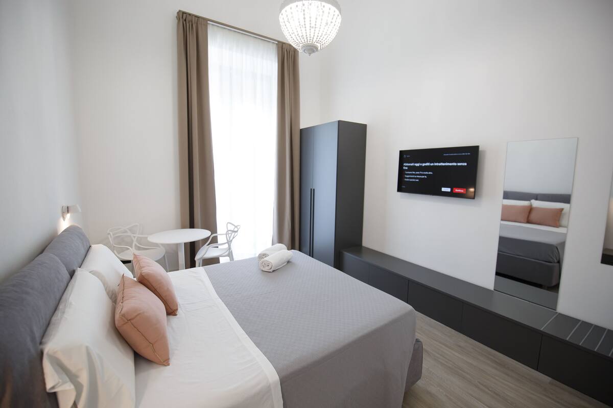 A modern hotel room with a neatly made bed, a television on the wall, a small table with chairs, and a wardrobe.