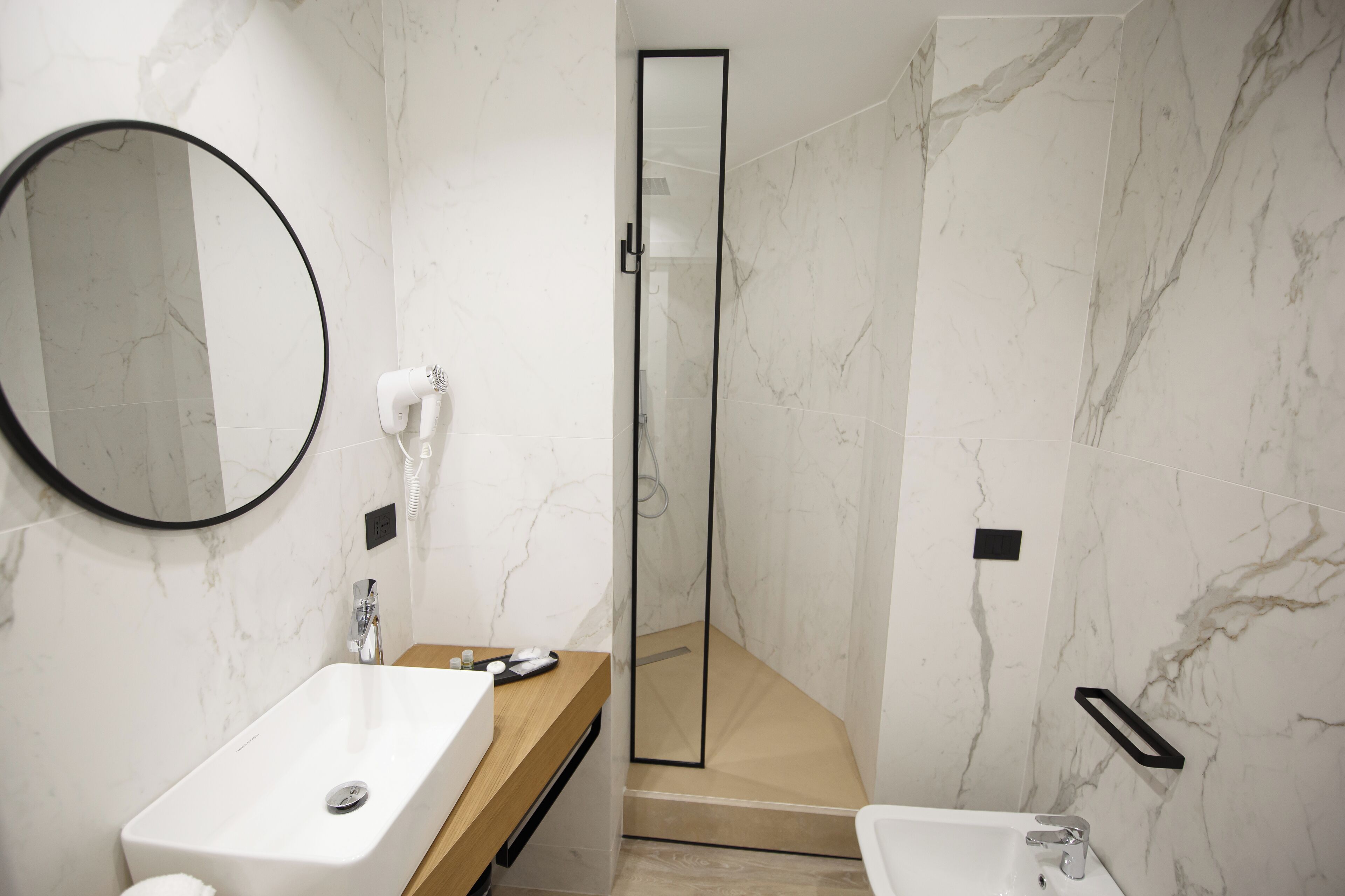 A modern bathroom with a marble wall, a large round mirror, a wooden vanity, a white sink, a glass shower enclosure, and a separate white pedestal sink.