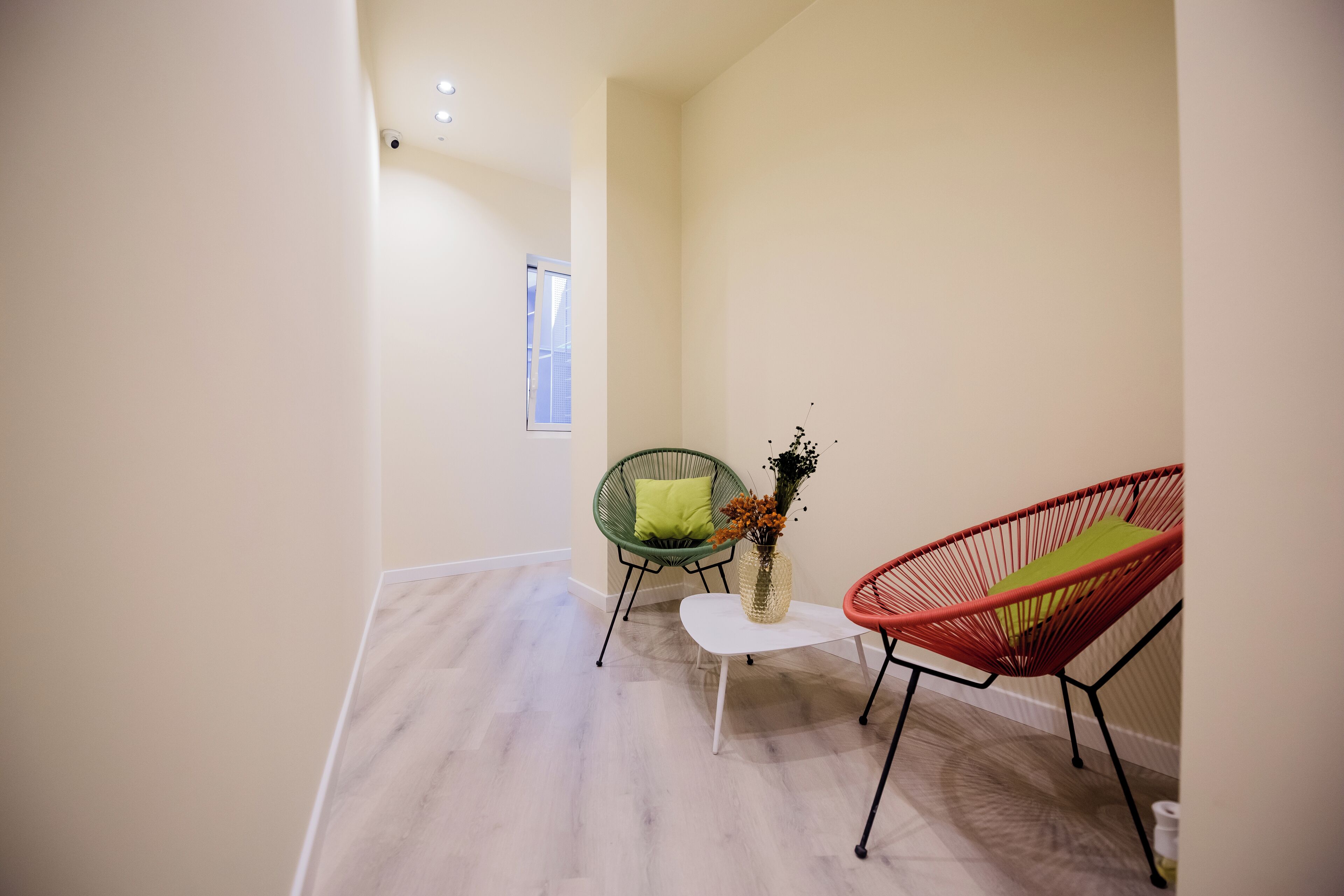 A modern room with a light wooden floor, a white table, two chairs with green and red cushions, and a vase with flowers.