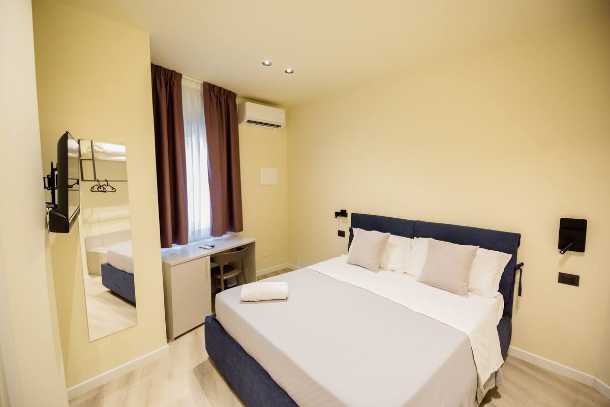 A hotel room with a bed, a desk, a mirror, an air conditioning unit, and a window with curtains.