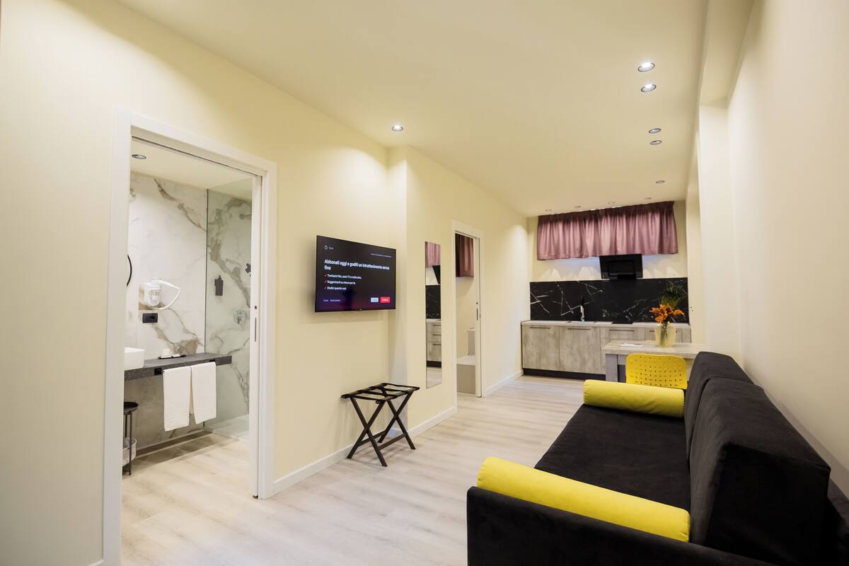 A modern living room with a black and yellow sofa, a flat-screen TV mounted on the wall, and a kitchen area in the background.