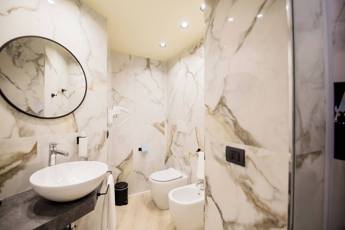 A modern bathroom with a marble wall, a round mirror, a white sink, a toilet, and a bidet.