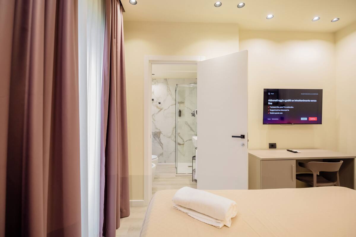 A modern hotel room with a bed, a desk, a TV, and a bathroom visible through an open door.