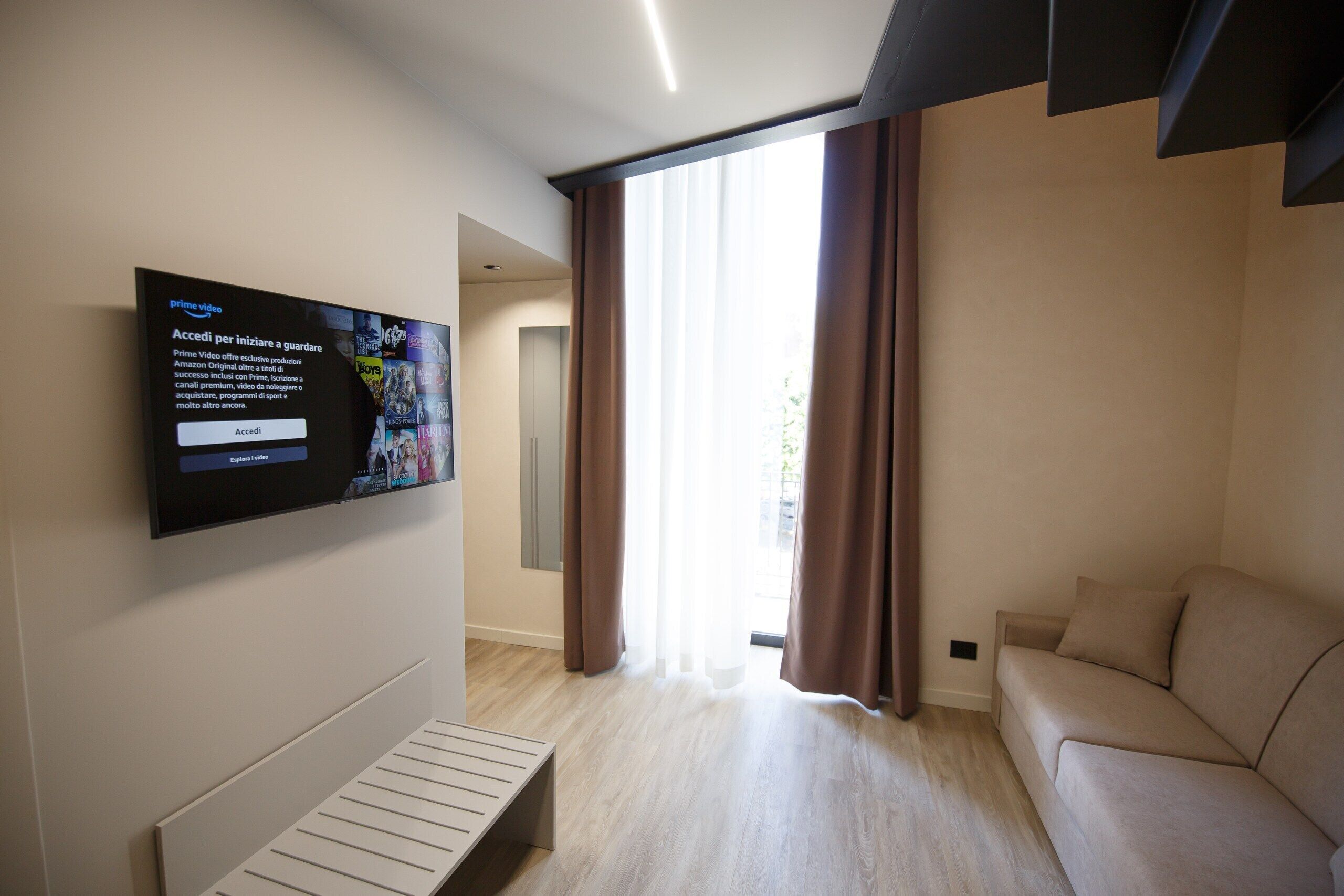 Un salon moderne doté d’un téléviseur à écran plat fixé au mur, d’un canapé, d’un banc et d’une grande fenêtre avec des rideaux.