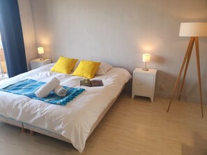 Una cama bien hecha con sábanas blancas y dos almohadas amarillas brillantes, una manta azul y una toalla enrollada. Hay dos mesitas de noche con lámparas, una a cada lado de la cama.