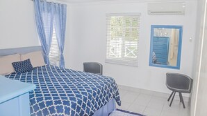 A bedroom with a bed, two chairs, a dresser, a window with blinds, and a mirror with a blue frame. - Bamboo Villa  (St. Ann's Bay)