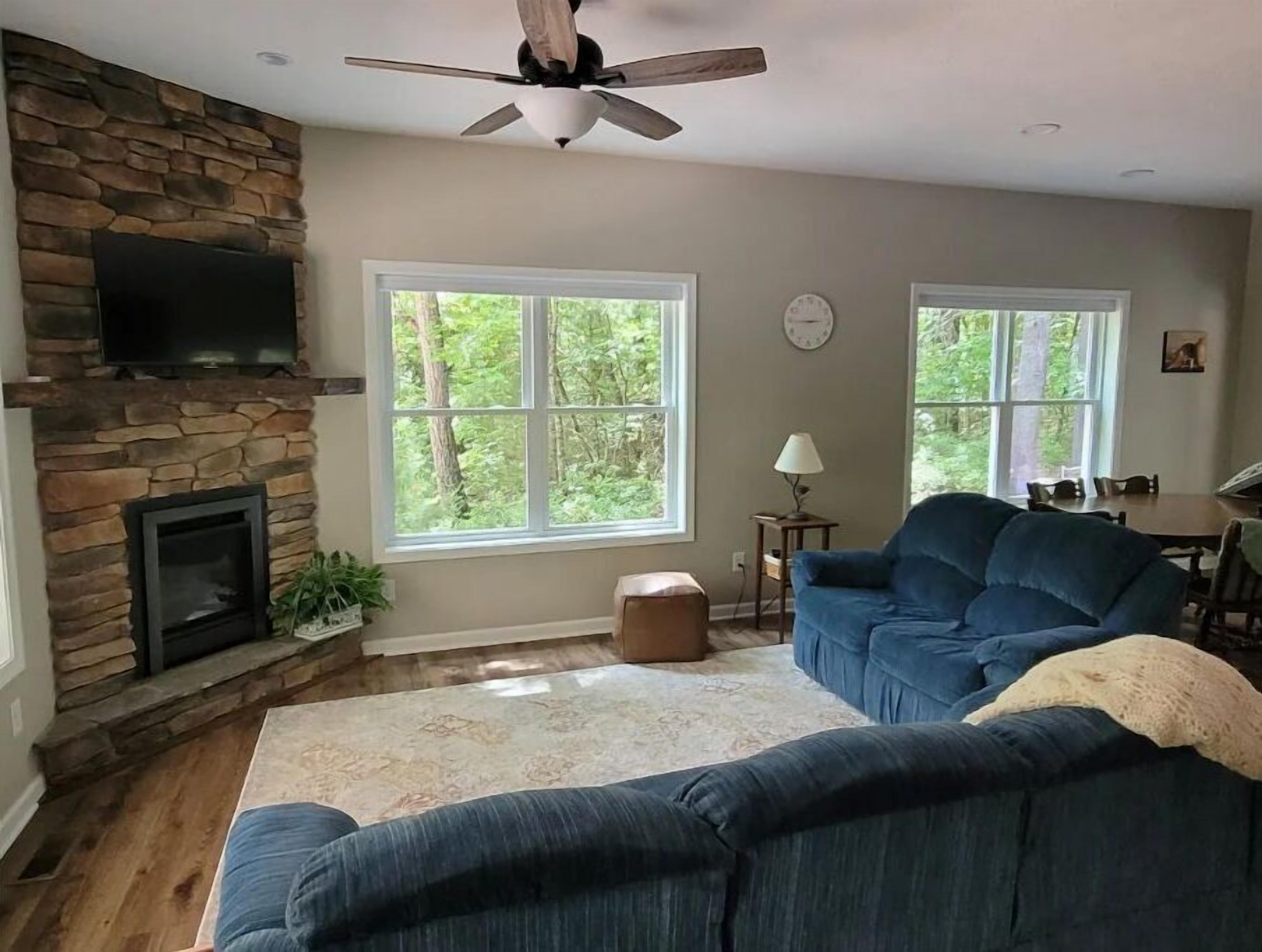 A living room with a stone fireplace, a ceiling fan, and large windows.