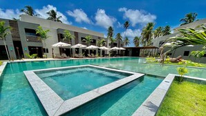 A swimming pool with a geometric tile design, surrounded by lounge chairs and umbrellas, with a modern building and palm trees in the background. - Villas Manatee by ER (Porto de Pedras)