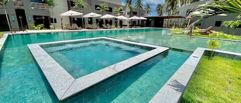 A swimming pool with a geometric tile design, surrounded by lounge chairs and umbrellas, with a modern building and palm trees in the background.