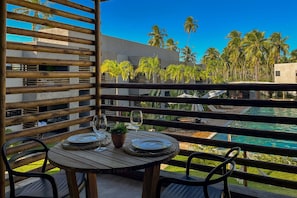 A balcony with a wooden table set for two, overlooking a swimming pool and palm trees. - Villas Manatee by ER (Porto de Pedras)