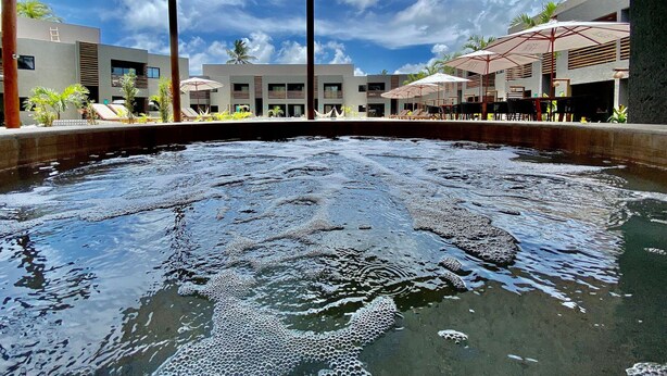 A swimming pool with a wooden canopy overhead, surrounded by modern buildings and outdoor seating areas. - Villas Manatee by ER (Porto de Pedras)