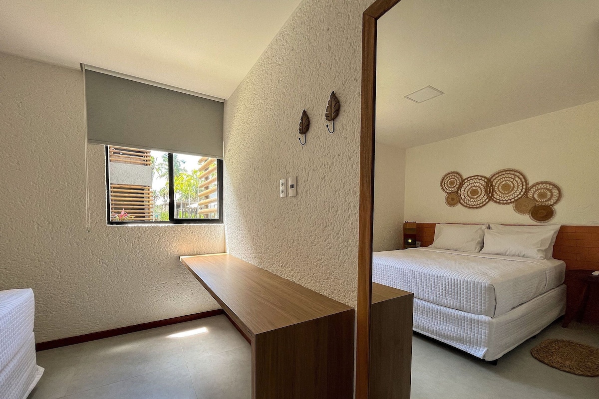 A hotel room with a bed, a wooden bench, a small table, a window with blinds, and a wall-mounted decorative piece.