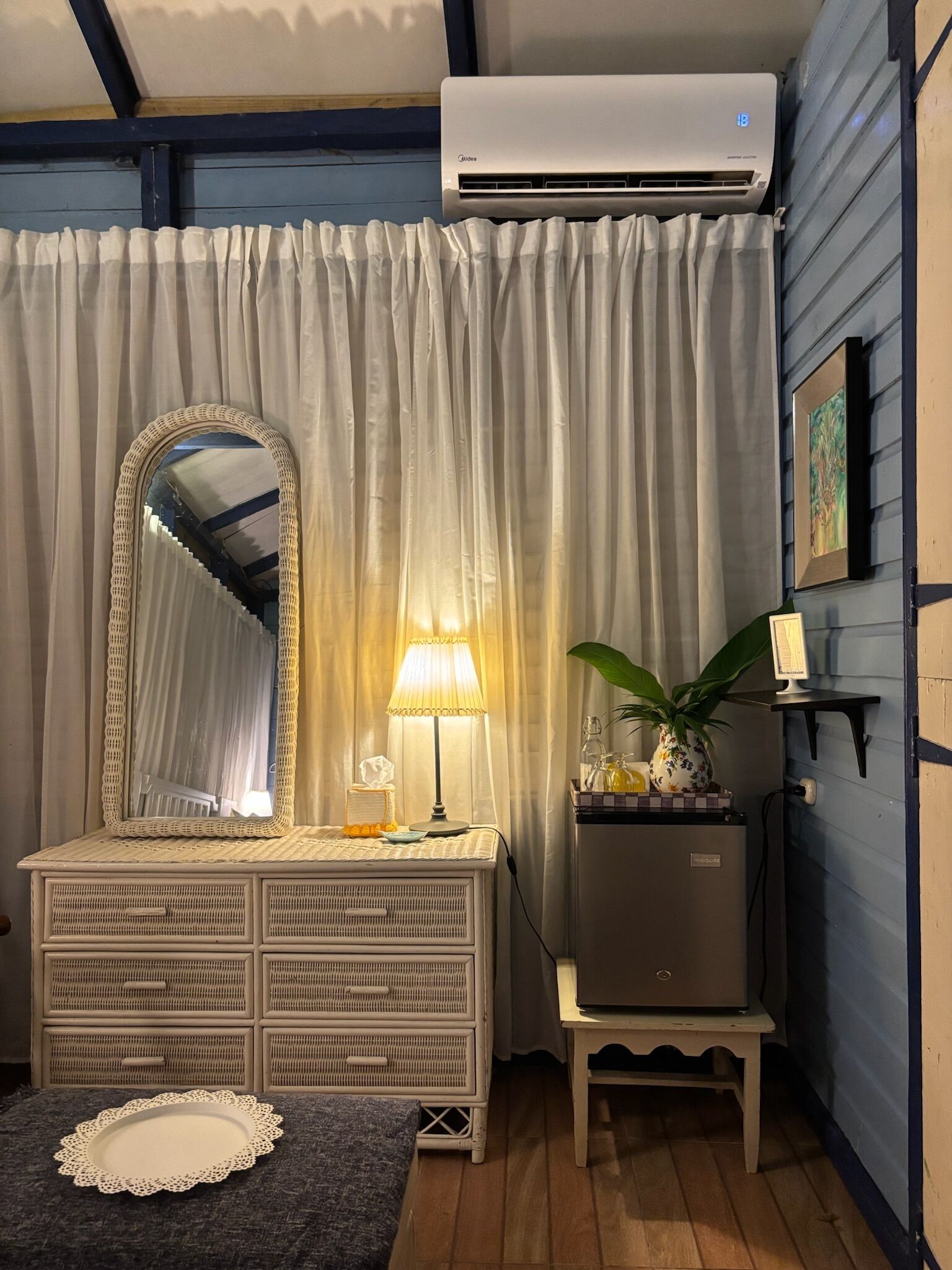 Quarto com cômoda branca, espelho, luminária, mesinha com planta e ar-condicionado.
