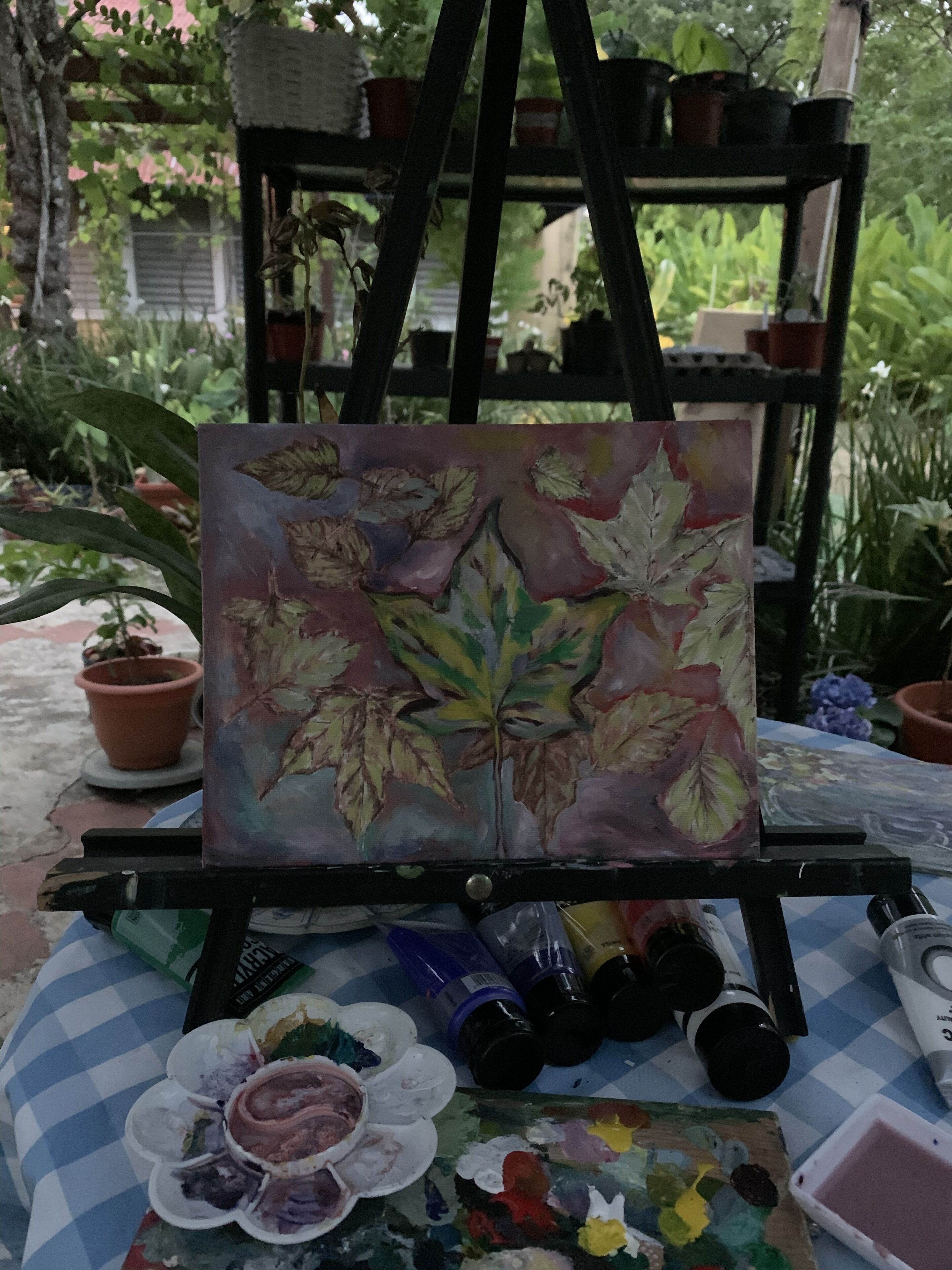 Een schilderstatief met een schilderij van bladeren, omringd door kunstbenodigdheden en planten in potten.