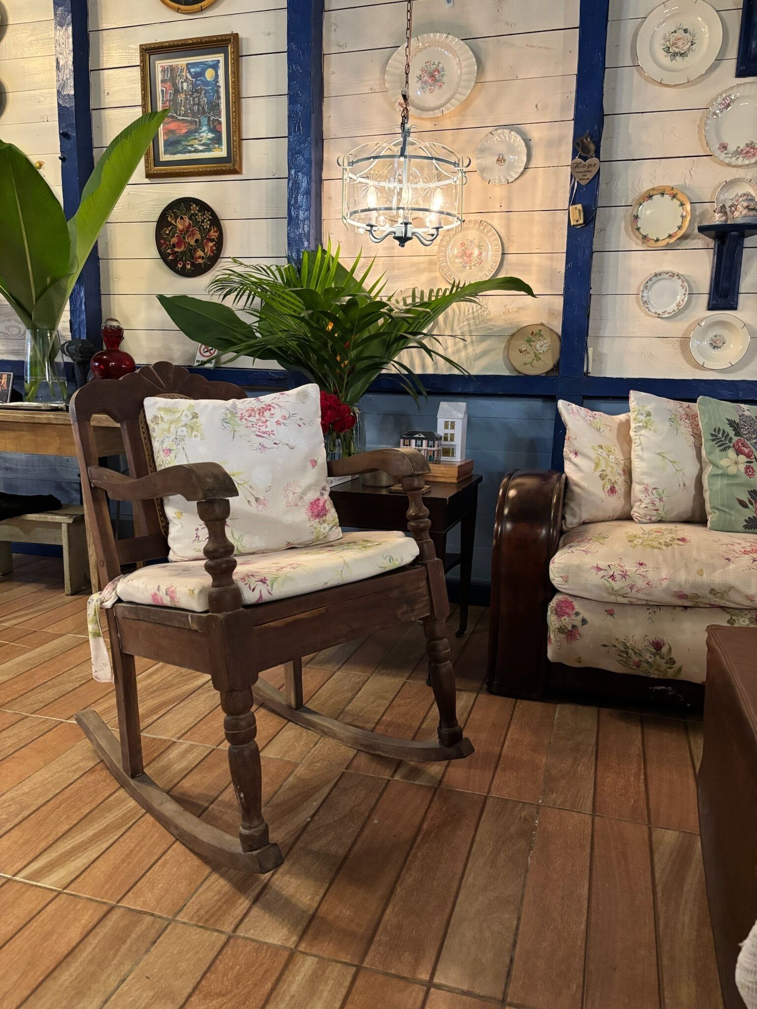 A room with a wooden rocking chair, a floral sofa, a chandelier, and wall-mounted plates.
