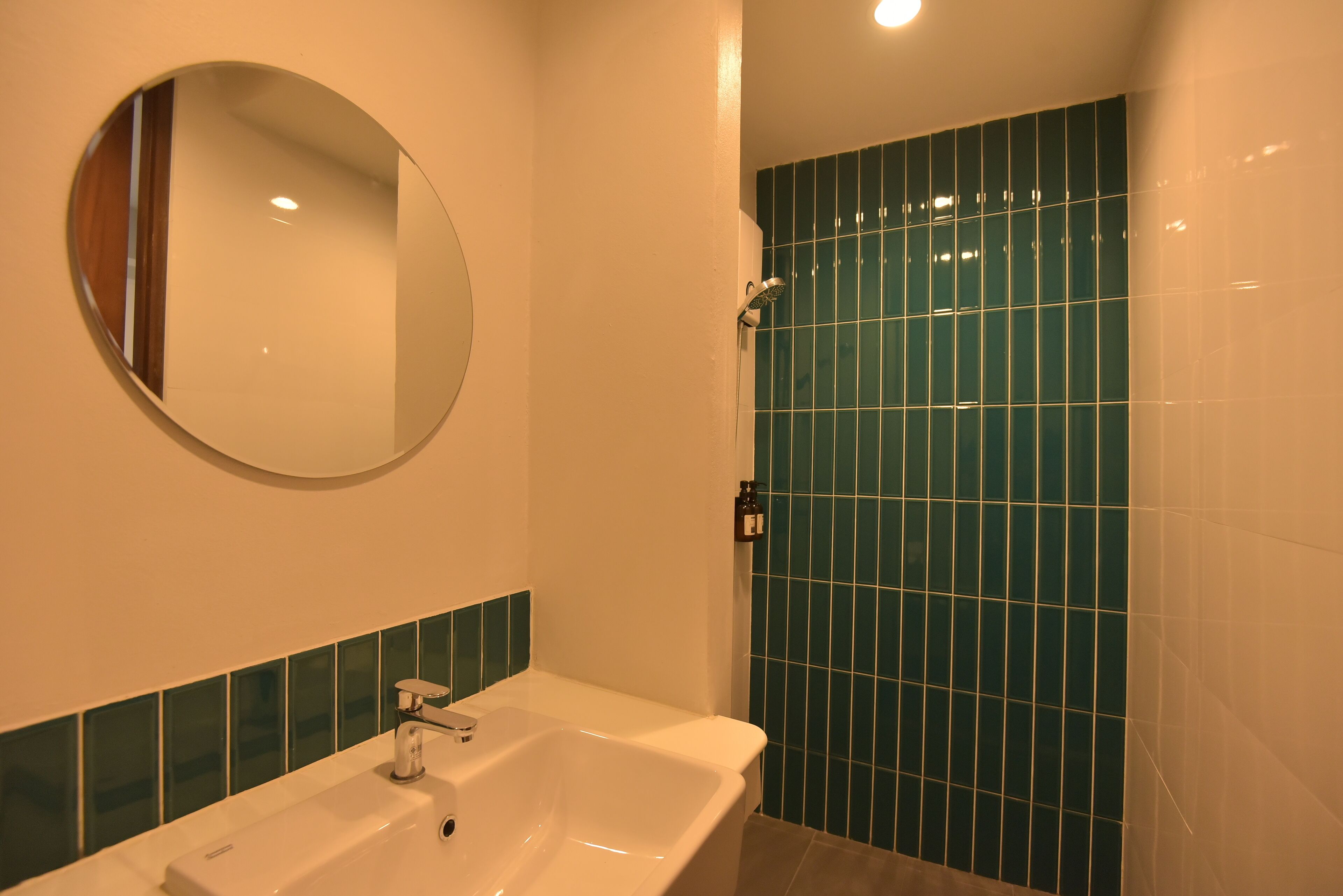 Ein Badezimmer mit weißem Waschbecken, einem runden Spiegel und einer Dusche mit grünen Fliesen.
