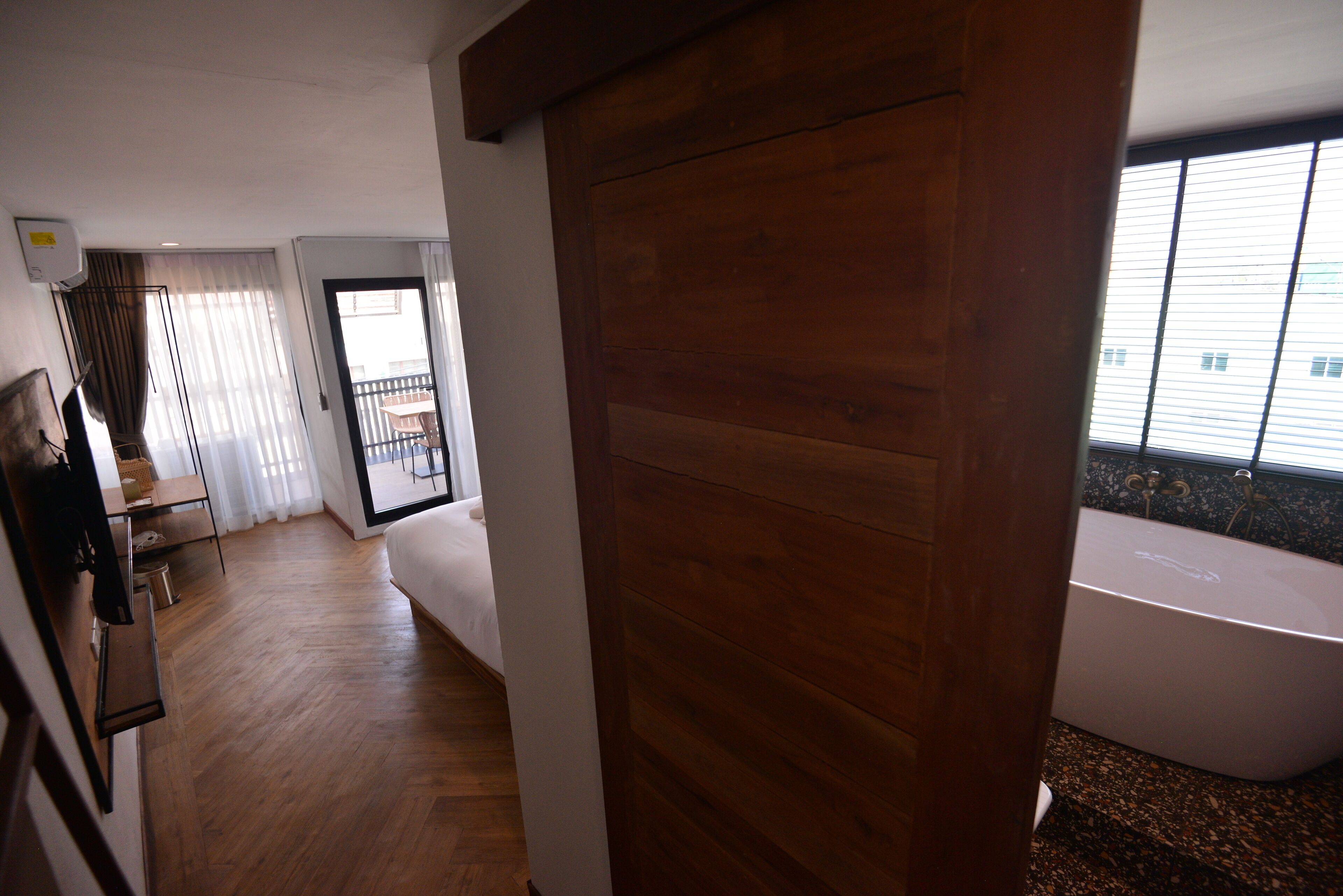 Une chambre d’hôtel avec un plancher en bois, une grande fenêtre avec des stores, une baignoire blanche et un balcon avec une table et des chaises.