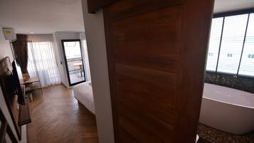 Une chambre d’hôtel avec un plancher en bois, une grande fenêtre avec des stores, une baignoire blanche et un balcon avec une table et des chaises.