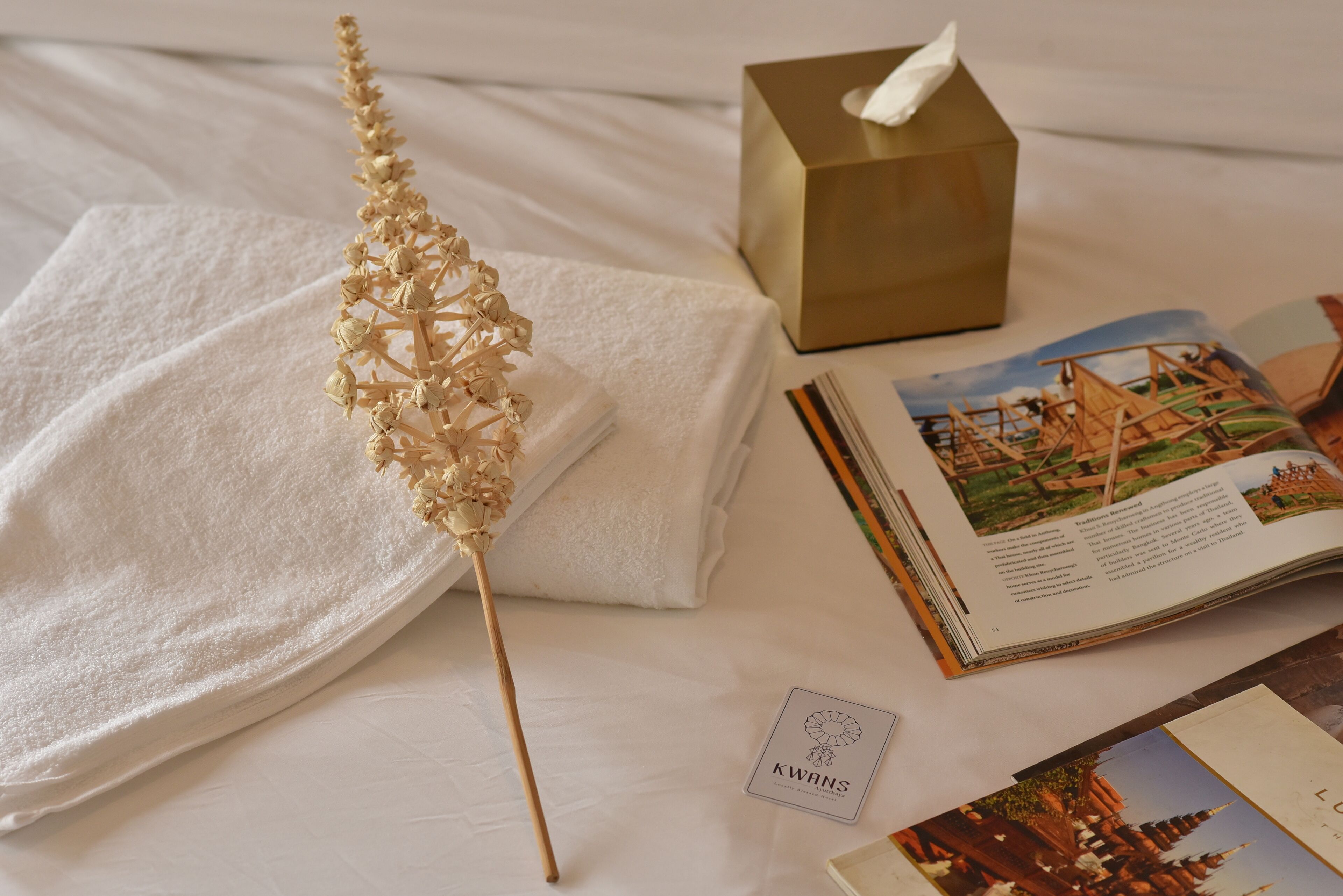 Una cama ordenada con una toalla blanca, una planta seca decorativa, una caja de pañuelos y un libro con una ilustración de viajes.
