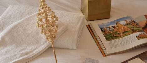A neatly arranged bed with a white towel, a decorative dried plant, a tissue box, and a book with a travel illustration.