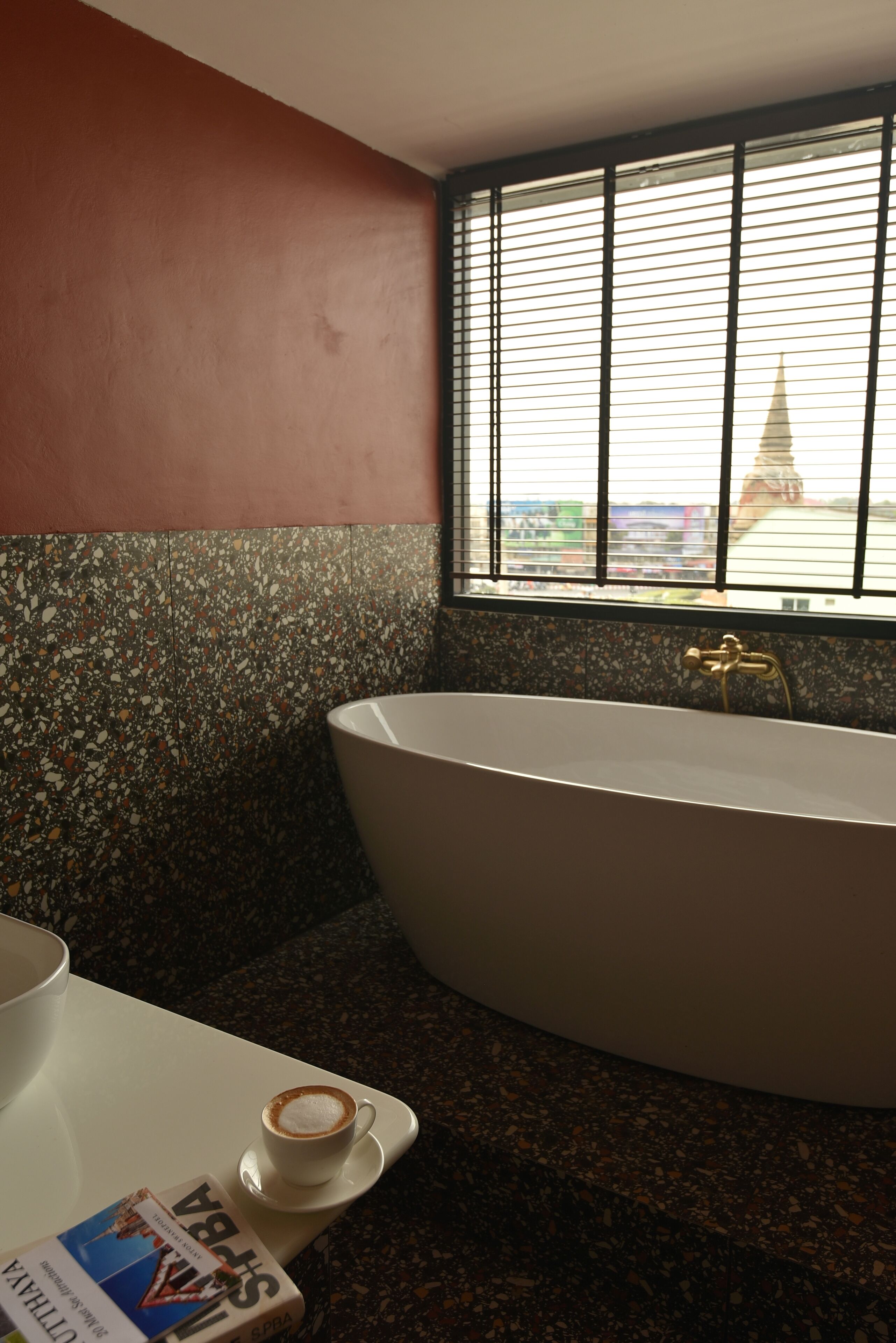 A bathroom with a freestanding bathtub, a window with blinds, a white sink, and a cup of coffee on a tray.