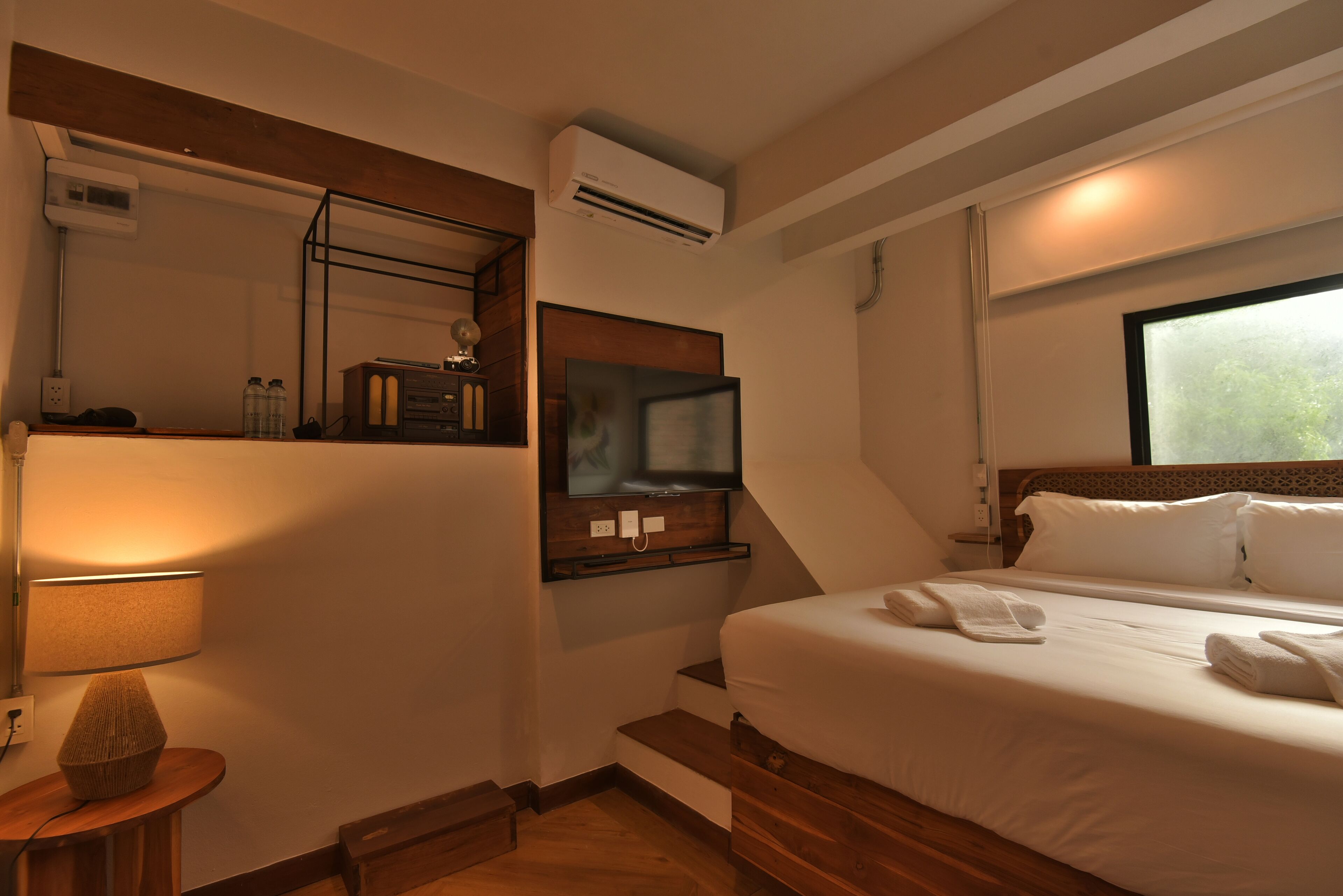 A hotel room with a bed, a TV mounted on the wall, a lamp, and a window.