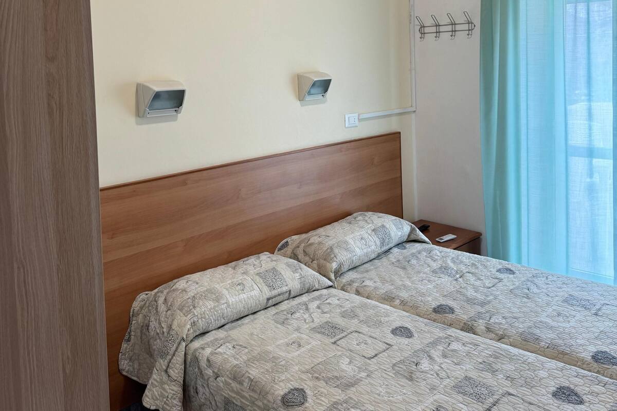 A hotel room with a wooden headboard, two beds with patterned bedding, an air conditioning unit, a small bedside table, a hook on the wall, and a window with light blue curtains.
