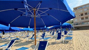 A beach with rows of blue lounge chairs and umbrellas, a sandy beach, and a building in the background. - Hotel Valente (Ortona)