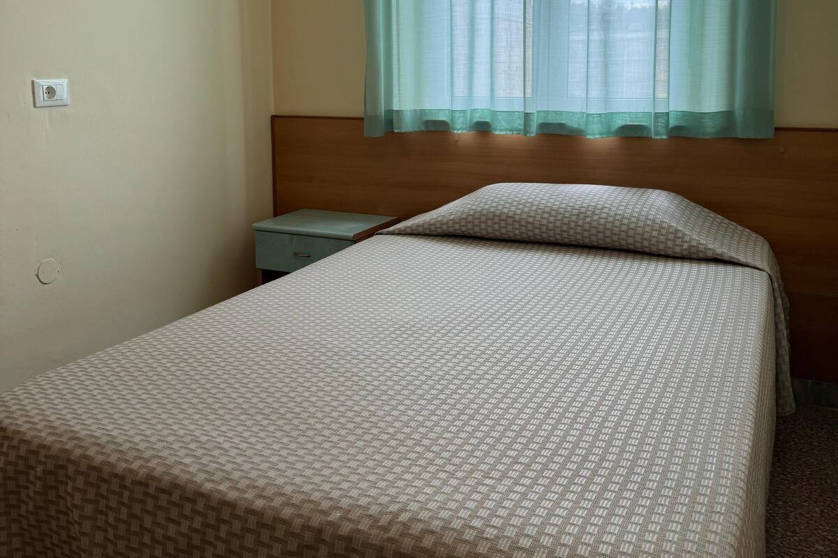 A room with a bed, a nightstand, a window with a sheer curtain, and a wall socket.