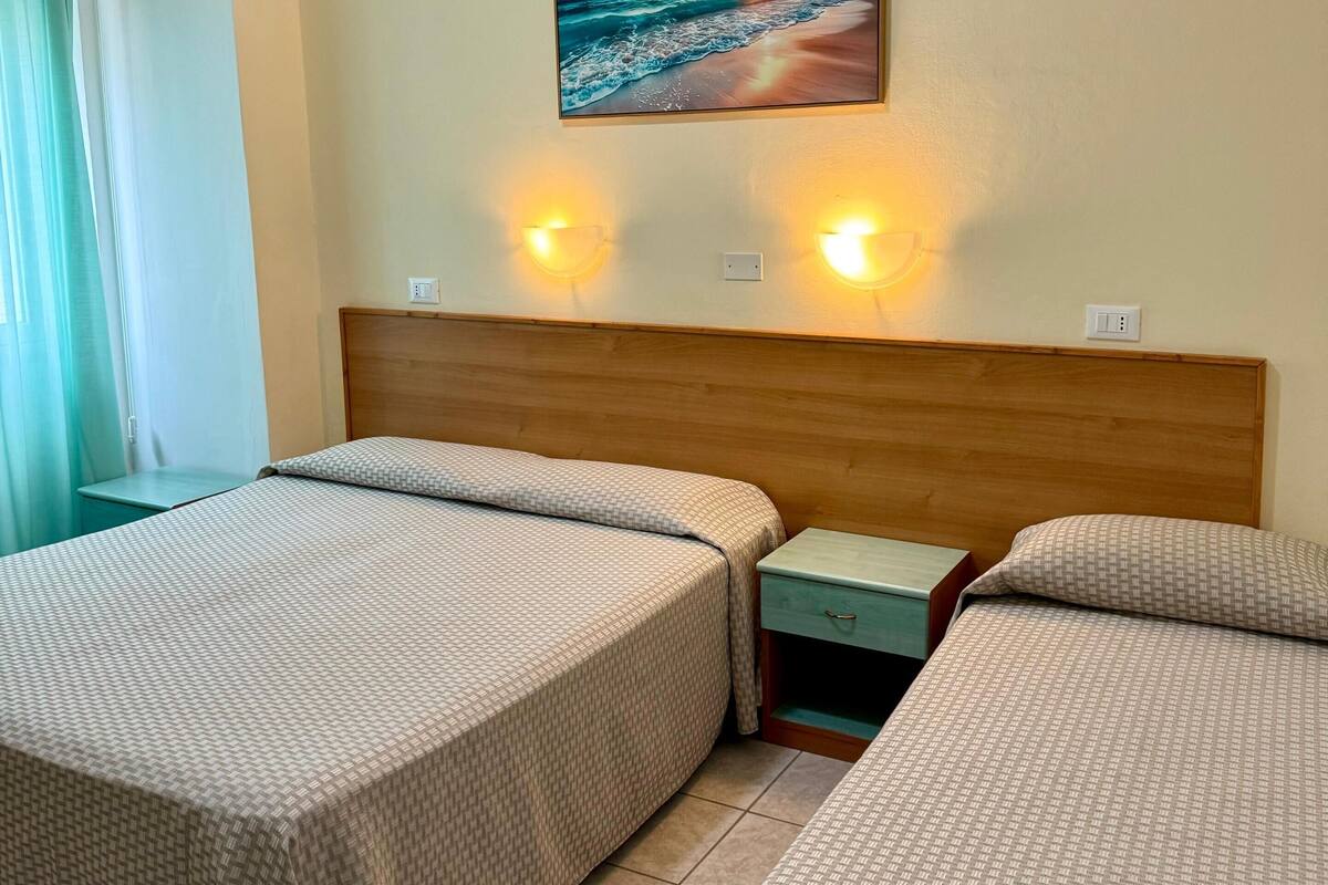 A hotel room with two beds, a wooden headboard, a painting of a beach scene, and two wall-mounted lights.