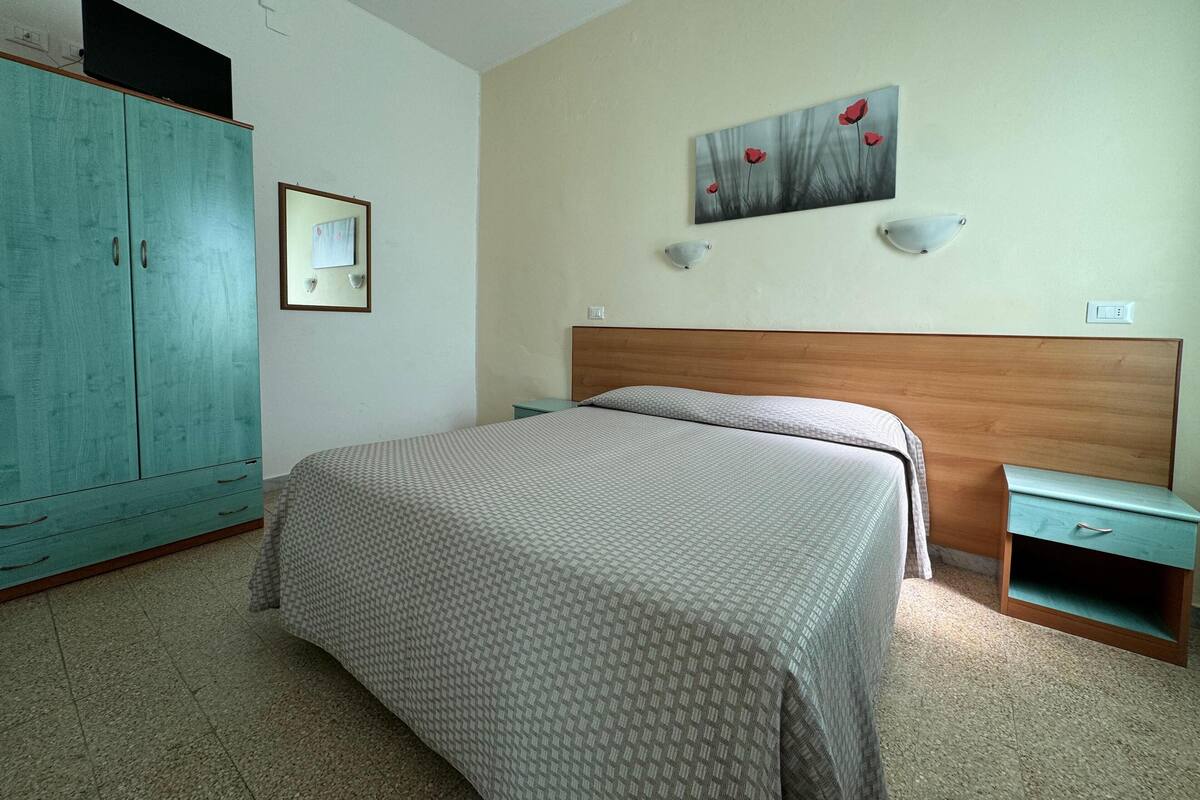 A bedroom with a bed, a wardrobe, a nightstand, a wall-mounted lamp, and a painting.