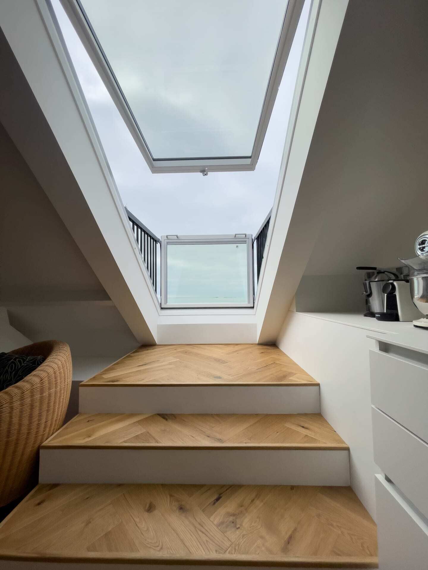 Une chambre à coucher de style loft moderne, dotée d’une lucarne, d’un escalier en bois, d’une commode blanche et d’un coin cuisine.