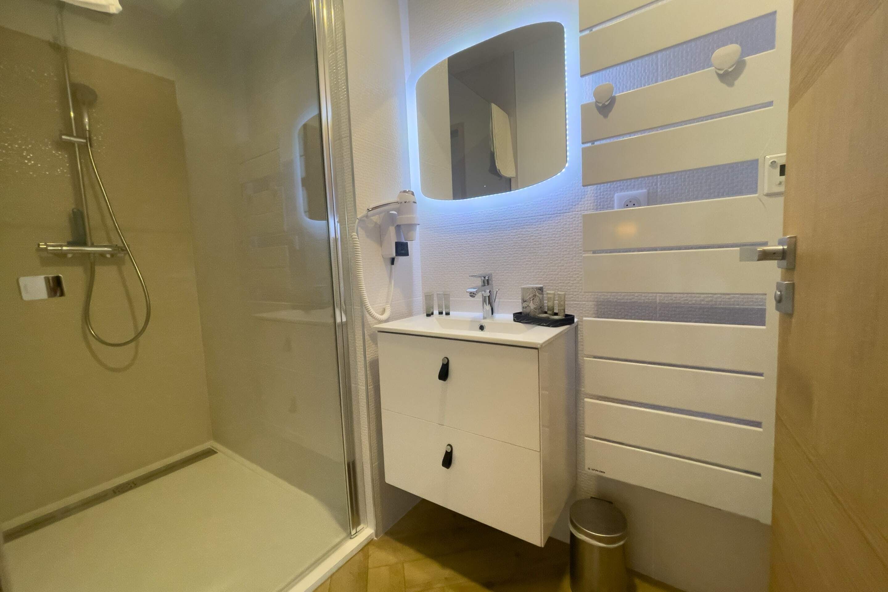 Ein modernes Badezimmer mit einer gläsernen Dusche, einem weißen Waschtisch mit Spiegel und einer kleinen Mülltonne.