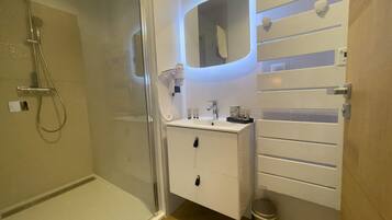 Ein modernes Badezimmer mit einer gläsernen Dusche, einem weißen Waschtisch mit Spiegel und einer kleinen Mülltonne.