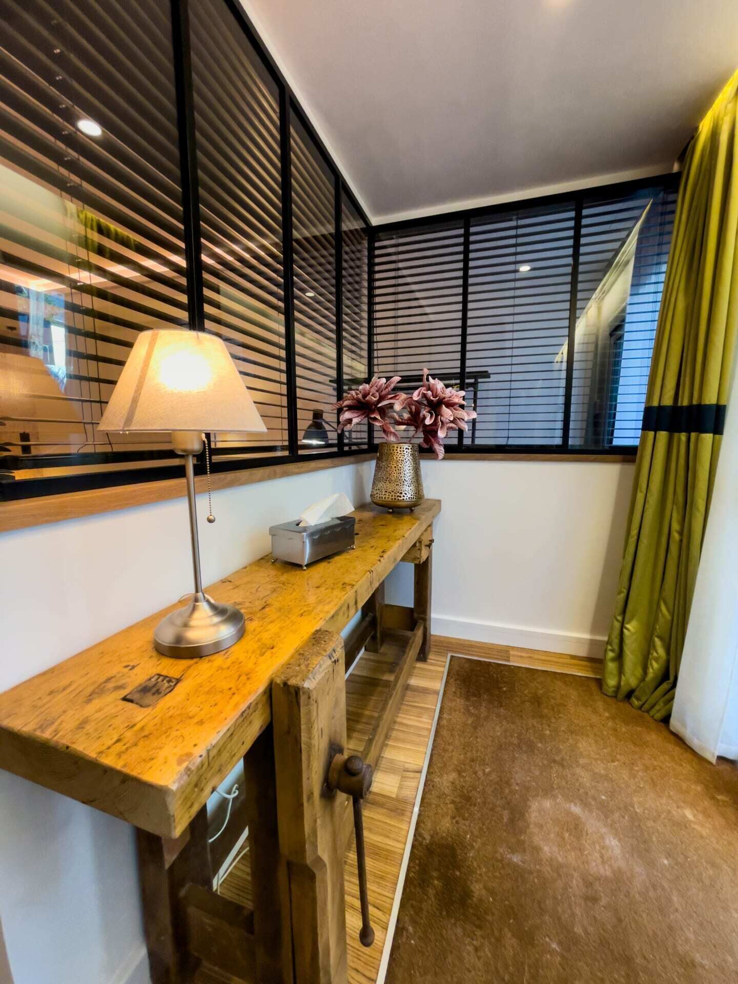 Una habitación con escritorio de madera, lámpara, un jarrón con flores y ventana con persianas.