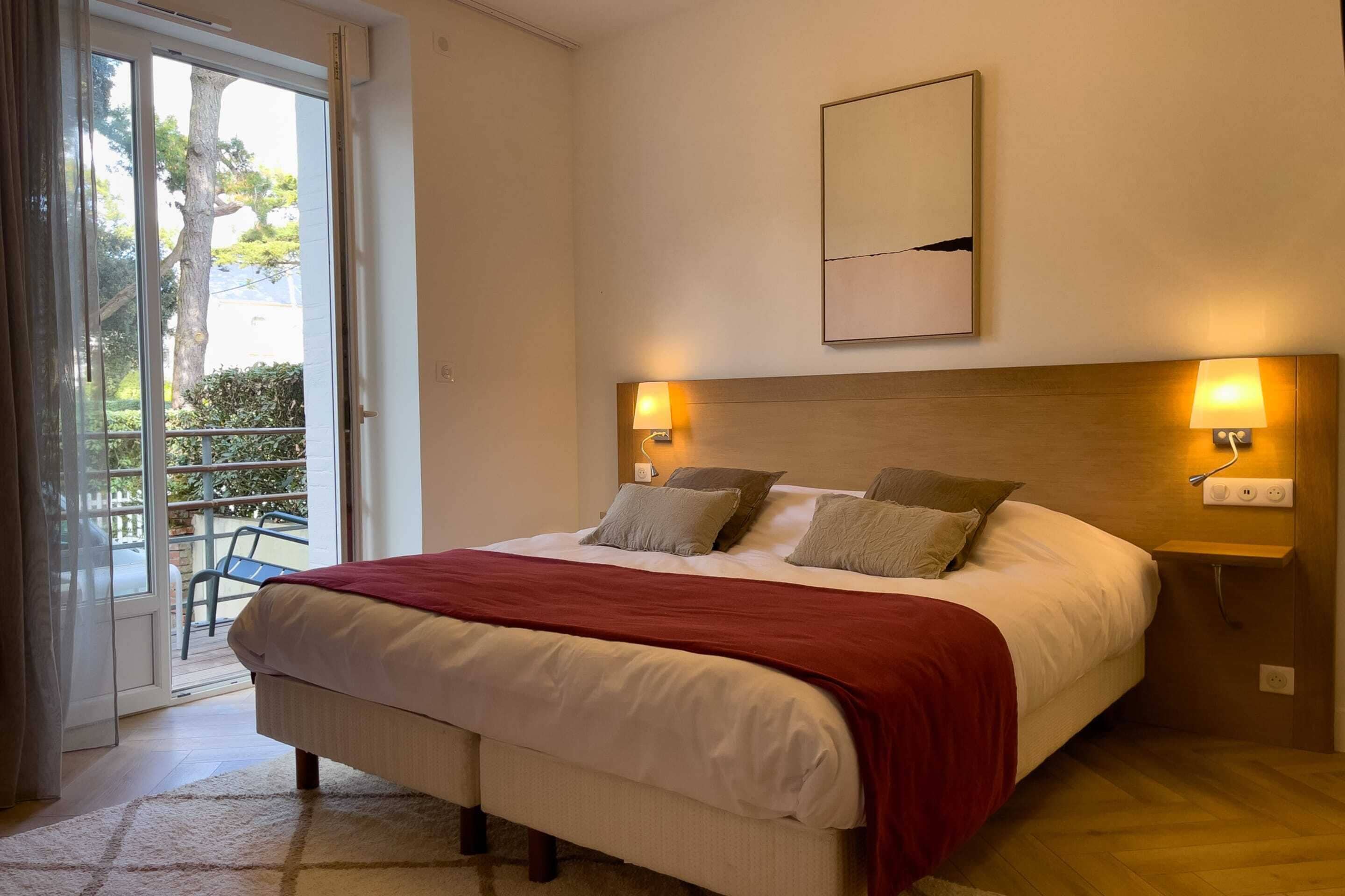 Une chambre dotée d’un grand lit, de deux lampes de chevet, d’une tête de lit en bois et donnant sur un espace extérieur grâce à une porte-fenêtre coulissante.