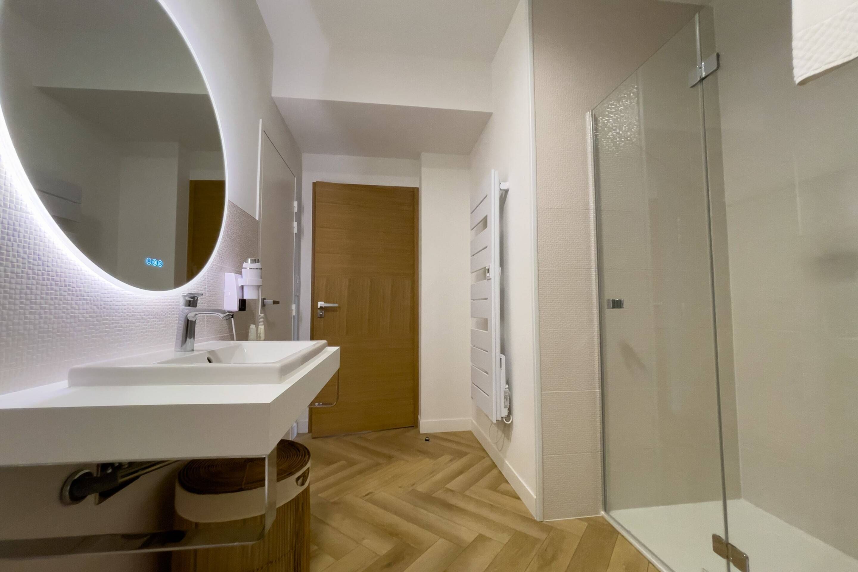 Ein modernes Badezimmer mit einem großen, ovalen Spiegel, einem weißen Waschbecken und einer Duschkabine aus Glas.