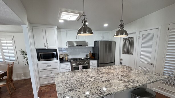 A modern kitchen with white cabinets, a granite island, stainless steel appliances, and pendant lighting.