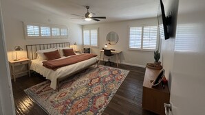 A bedroom with a bed, a desk, a chair, a ceiling fan, and a TV mounted on the wall.
