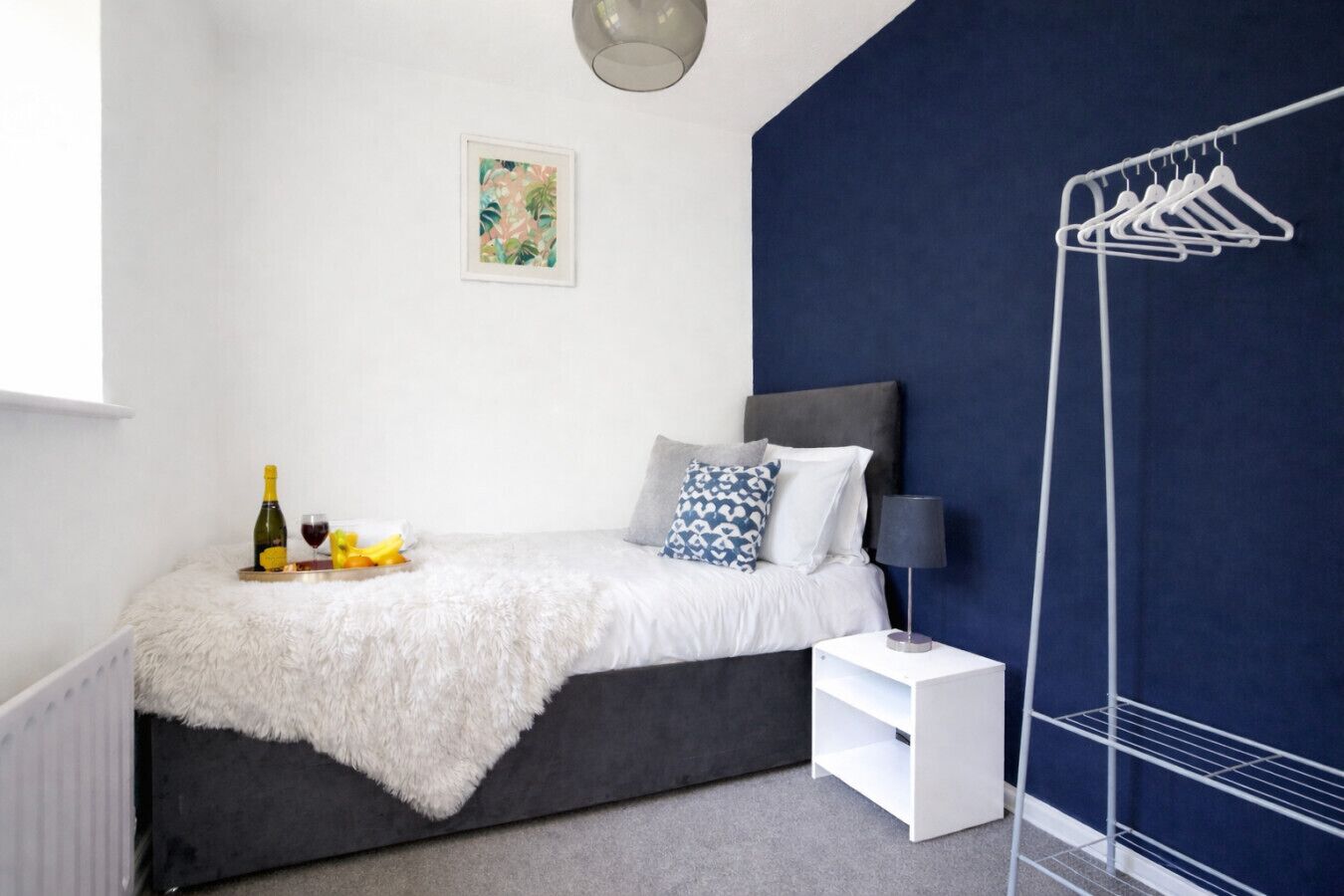 A bedroom with a bed, a bedside table, a lamp, a radiator, and a clothing rack.