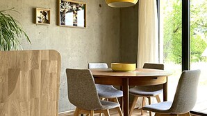 A dining area with a round wooden table, six chairs, a yellow pendant light, and a large window with a view of greenery. - Appartement Contemporain Avec Terrasse à 5mn de du lac D'annecy (Doussard)