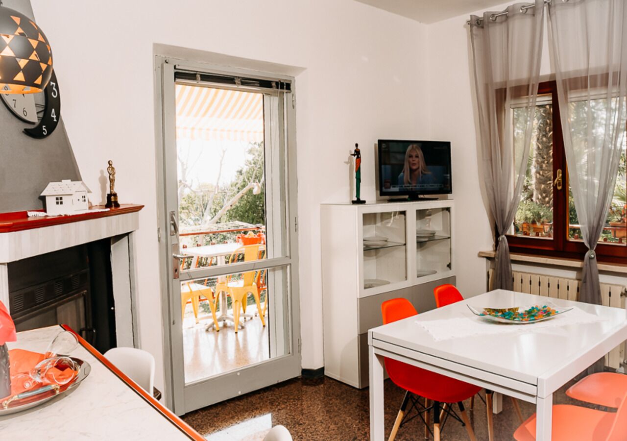 Ein modernes Wohnzimmer mit Kamin, einem Essbereich mit orangefarbenen Stühlen, einem Fernseher auf einem weißen Schrank und einem Balkon mit Gartenmöbeln.