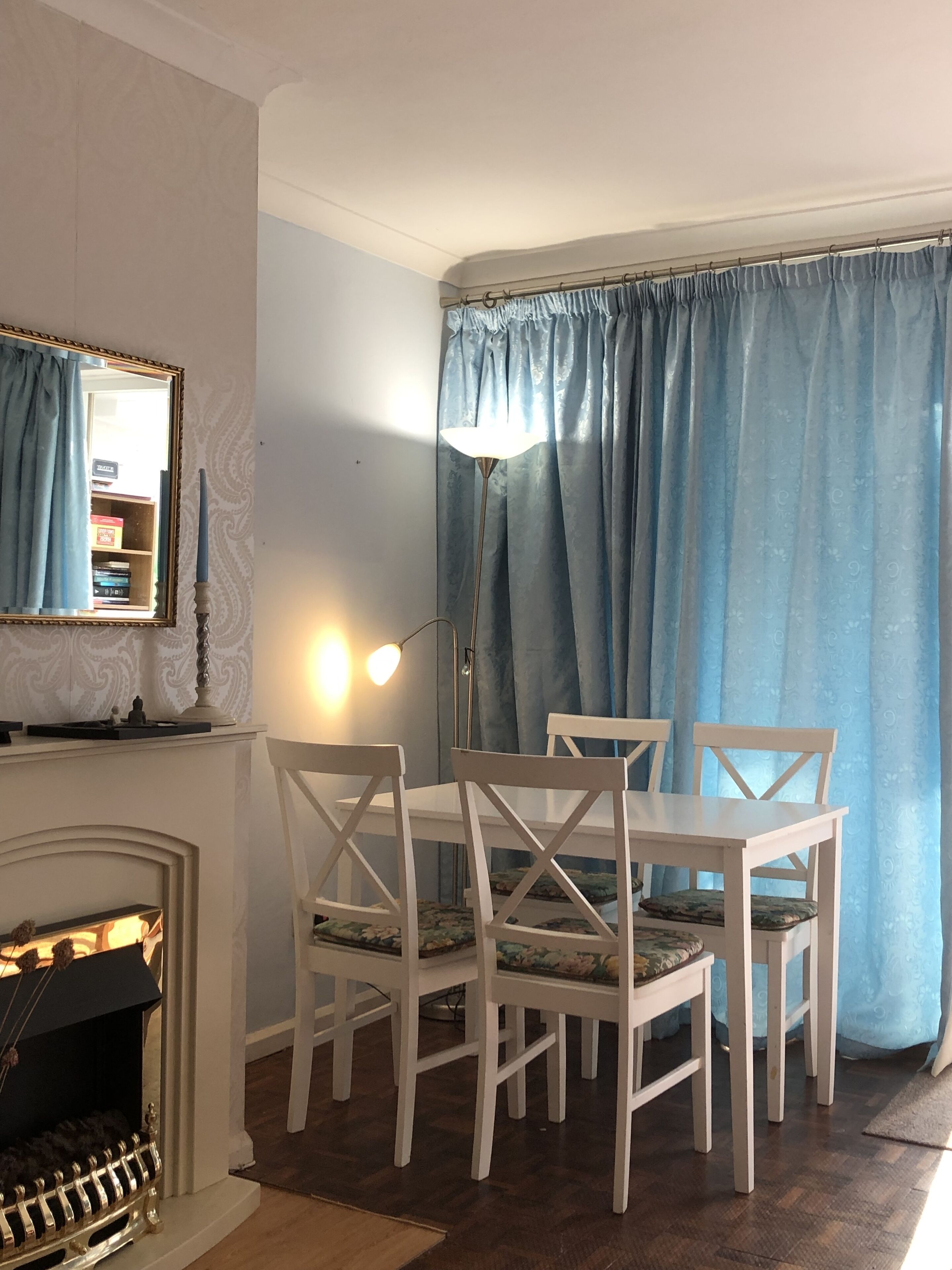 Un coin repas avec une table et des chaises blanches, une cheminée, un miroir et des rideaux bleus.