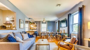 A modern living room with a sofa, coffee table, and chairs. There is a dining area with a table and chairs. The room has a ceiling fan, windows with curtains, and shelves with decorative items. - Pet-Friendly Pensacola Home w/ Sunny Backyard! (Pensacola)