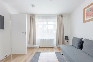 A room with a grey sofa, a white door, a window with curtains, and a radiator.