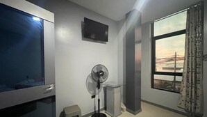 Een kamer met een televisie, een ventilator, een klein krukje en een raam met uitzicht op een brug.