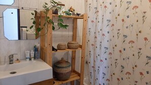 A bathroom with a wooden shelf holding baskets and a potted plant, a sink with toiletries, and a floral shower curtain. - Arlozorov 3BD Apartment (Tel Aviv)