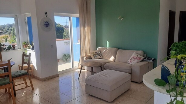 A living room with a beige sofa, a small table, and a chair. There is a balcony with plants and a view of greenery. - Arlozorov 3BD Apartment (Tel Aviv)