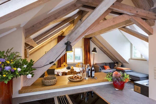 Une pièce mansardée chaleureuse avec des poutres apparentes, une table en bois agrémentée d’un panier et de fleurs, un canapé et une fenêtre avec des rideaux.