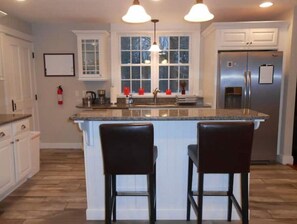 A kitchen with white cabinets, a central island with two bar stools, a stainless steel refrigerator, and pendant lighting. - Old World Charm, Modern Conveniences - 3 BR private home in quiet setting (Easthampton)